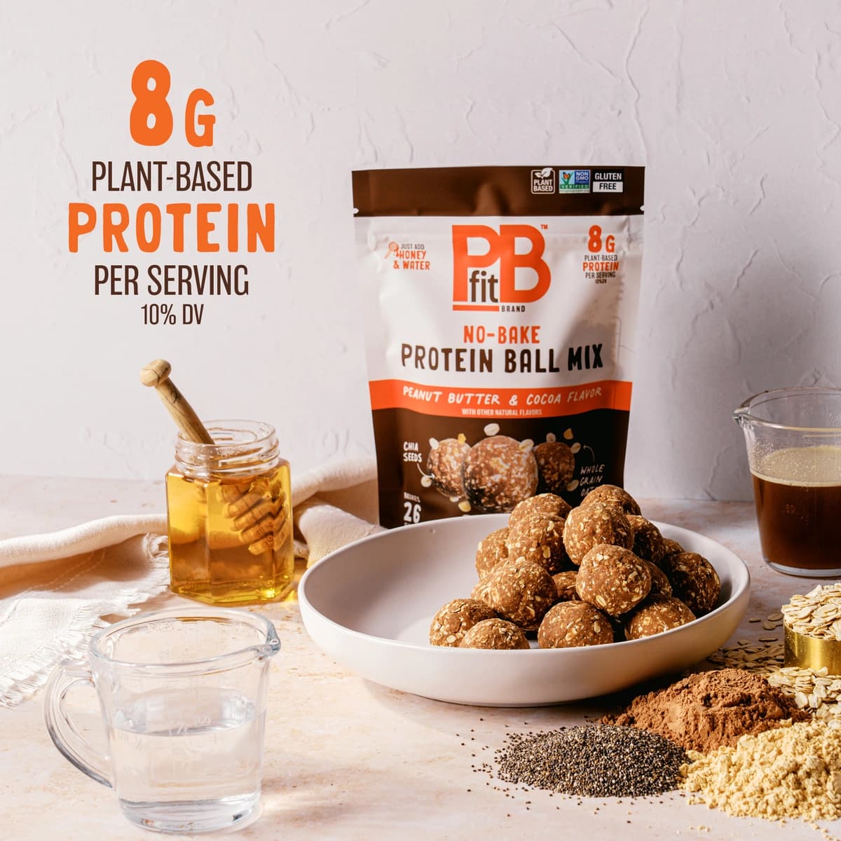 A pile of protein balls in a bowl next to the bowl is a jar of honey and a measuring cup of water as well as the ingredients that are in the PBfit Protein Ball Cocoa Mix