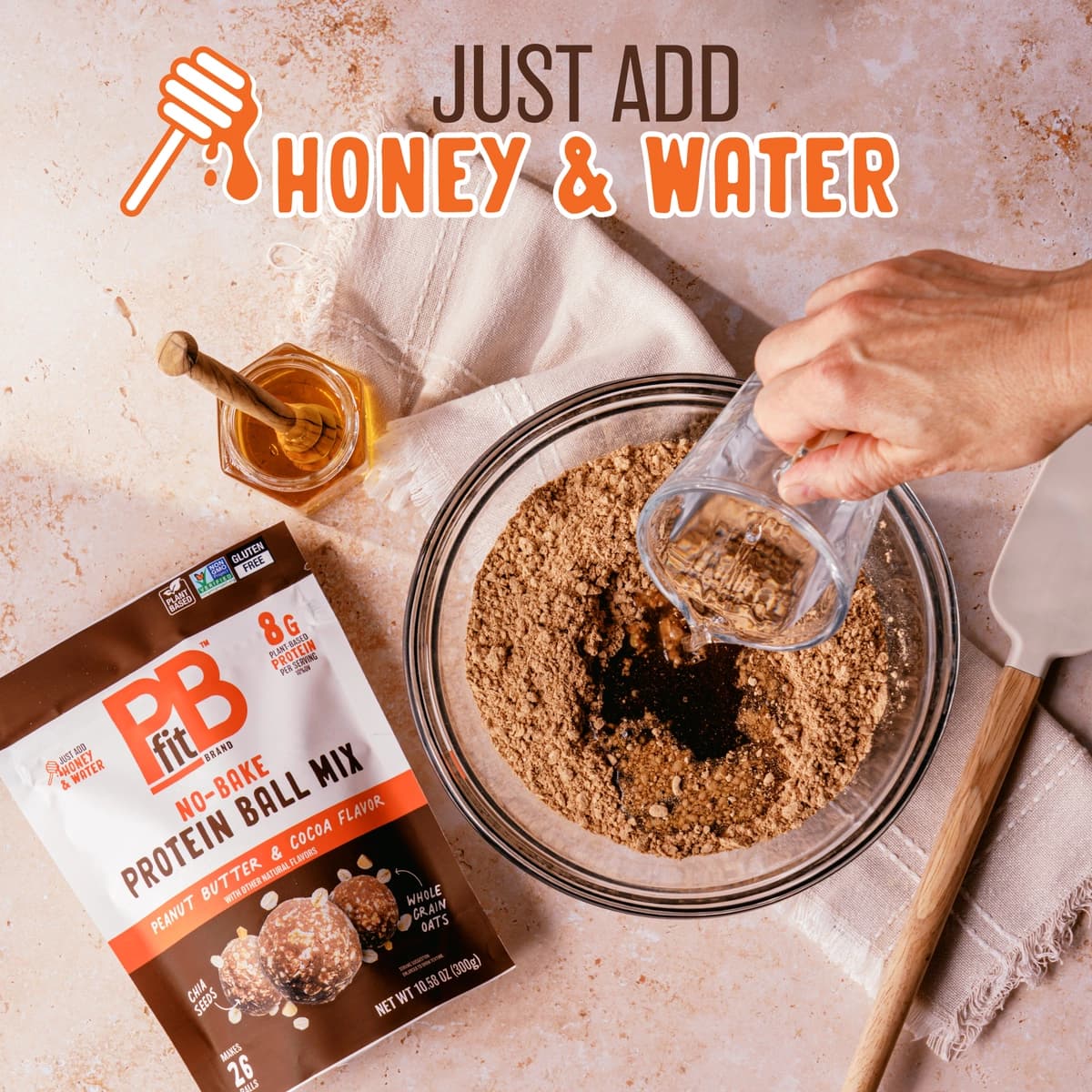 A person adding water into the PBfit Protein Ball Cocoa Mix in a bowl. Around the bowl there is a little jar of honey and a spoon to mix all the ingredients together.