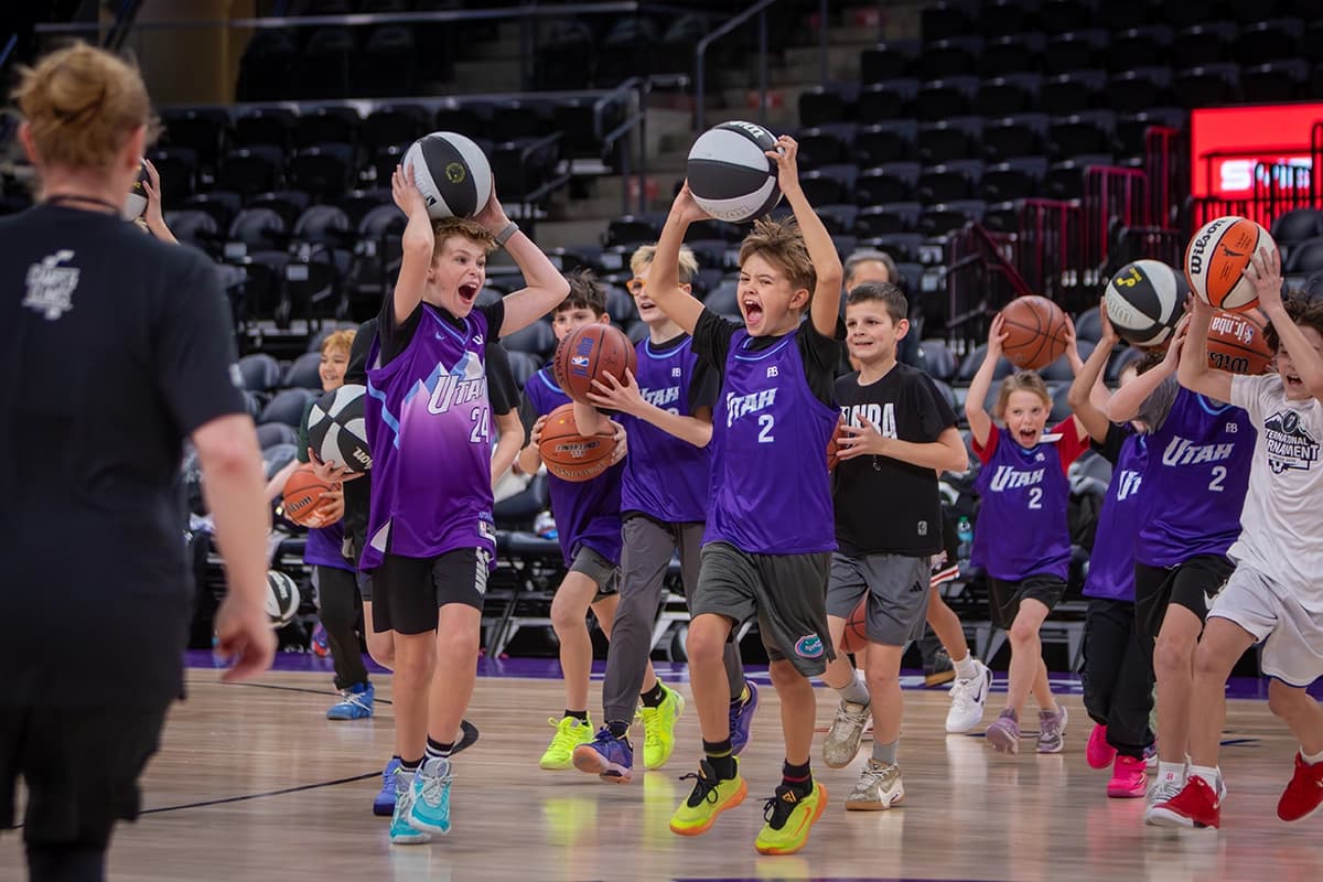 Young boy practicing basketball dribbling skills on an indoor court, wearing a jersey with the number 23. A blue banner in the background reads `Camps & Clinics presented by PBfit,` featuring a basketball and music note logo.