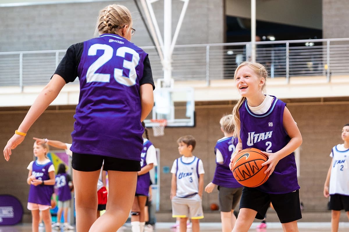 Young boy practicing basketball dribbling skills on an indoor court, wearing a jersey with the number 23. A blue banner in the background reads `Camps & Clinics presented by PBfit,` featuring a basketball and music note logo.